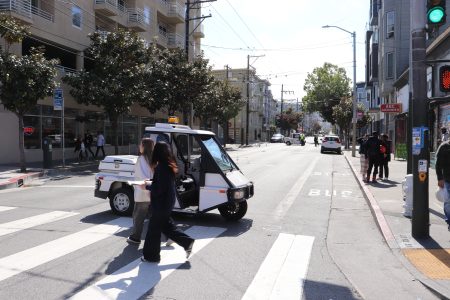 A small police vehicle is parked in the middle of a city crosswalk while pedestrians cross the street and the traffic light is green, illustrating concerns about SFMTA harassment.