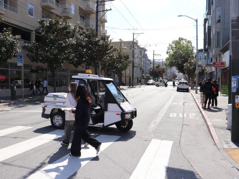 A small police vehicle is parked in the middle of a city crosswalk while pedestrians cross the street and the traffic light is green, illustrating concerns about SFMTA harassment.