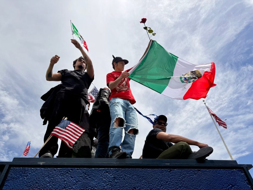 A group of people stand on a blue platform holding Mexican and American flags, with one person raising a rose. The sky is partly cloudy in the background.