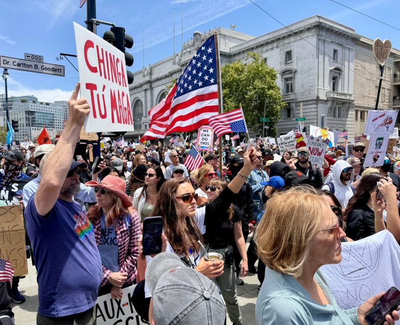 A large crowd of people protests outdoors, holding signs in different languages and American flags, with buildings and traffic lights visible in the background.