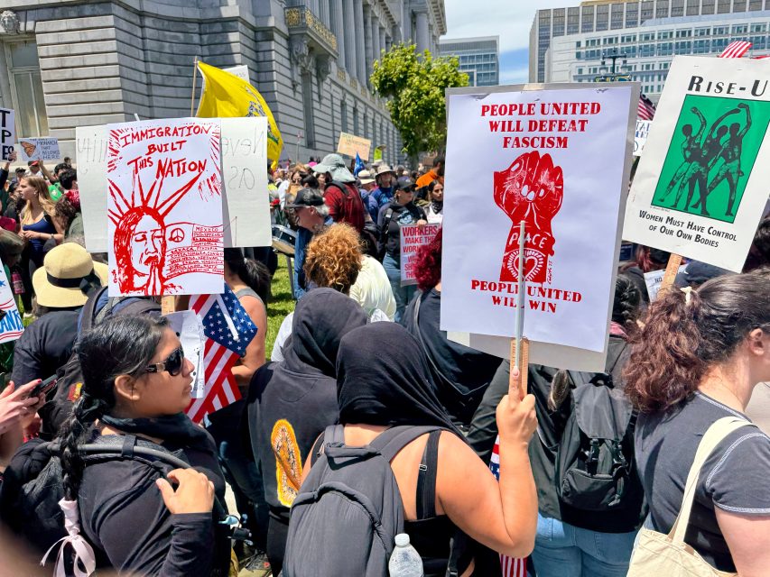 Protesters gathered outdoors holding signs with messages supporting immigration and anti-fascism, including “Immigration Built This Nation” and “People United Will Defeat Fascism.”.