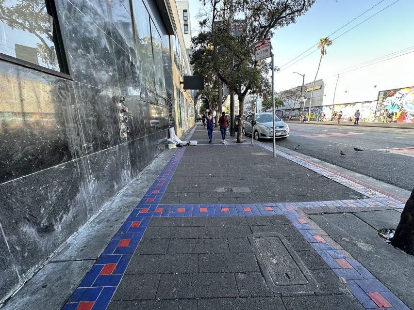 A city sidewalk with a few pedestrians, a car parked at the curb, some graffiti on distant walls, and a person sitting near a building entrance.