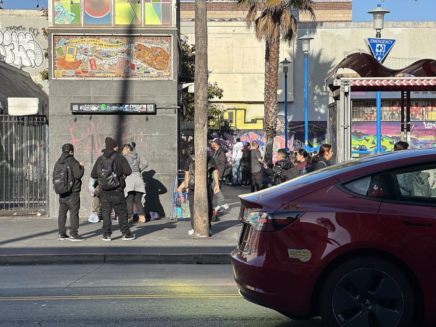 A group of people waits at a busy urban bus stop near a sidewalk with graffiti-covered walls; a red car is parked in the foreground.