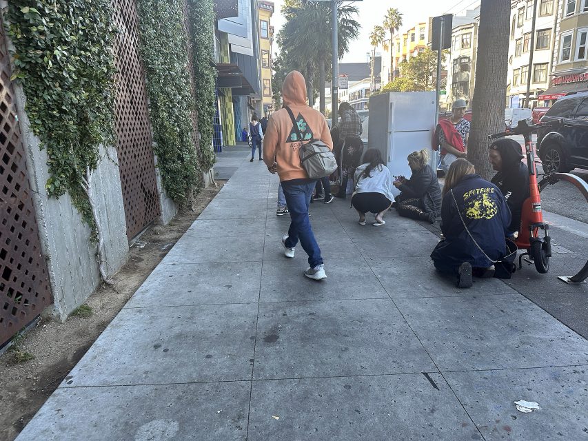 People are gathered on a city sidewalk near buildings and parked scooters; one person walks away wearing a tan hoodie and carrying a backpack.