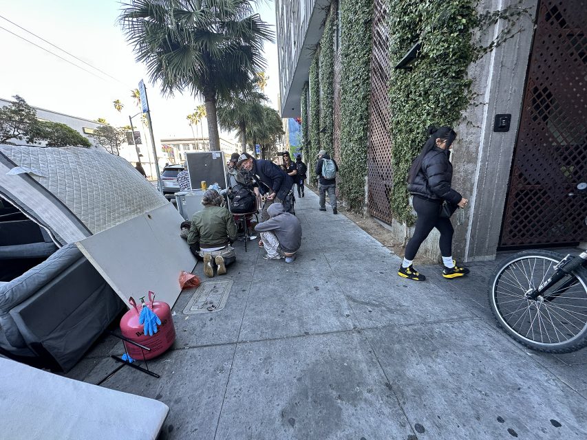 People interact on a city sidewalk next to tents and makeshift shelters, with pedestrians walking by and a bicycle partially visible in the corner.
