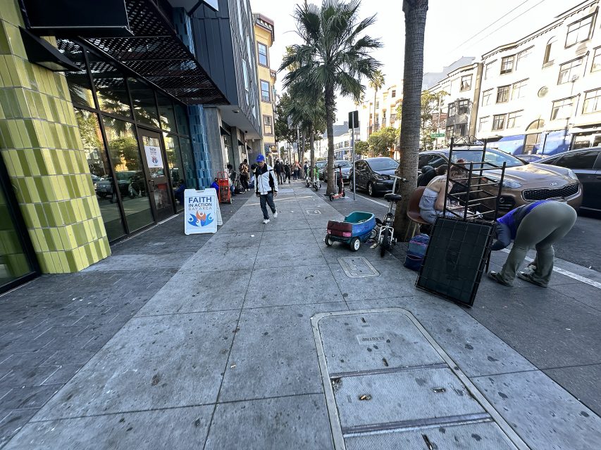 City sidewalk with pedestrians walking, a palm tree, and people organizing belongings near carts and bins; cars are parked along the street and buildings line both sides.