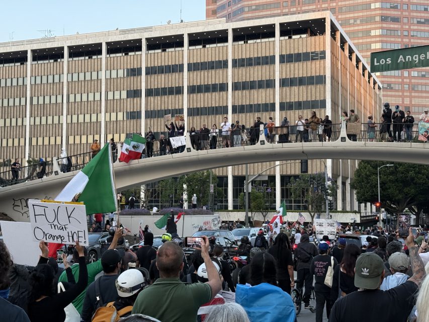 A crowd of protesters gathers on a city street and footbridge, holding signs, Mexican flags, and banners. A green street sign reads "Los Angeles.