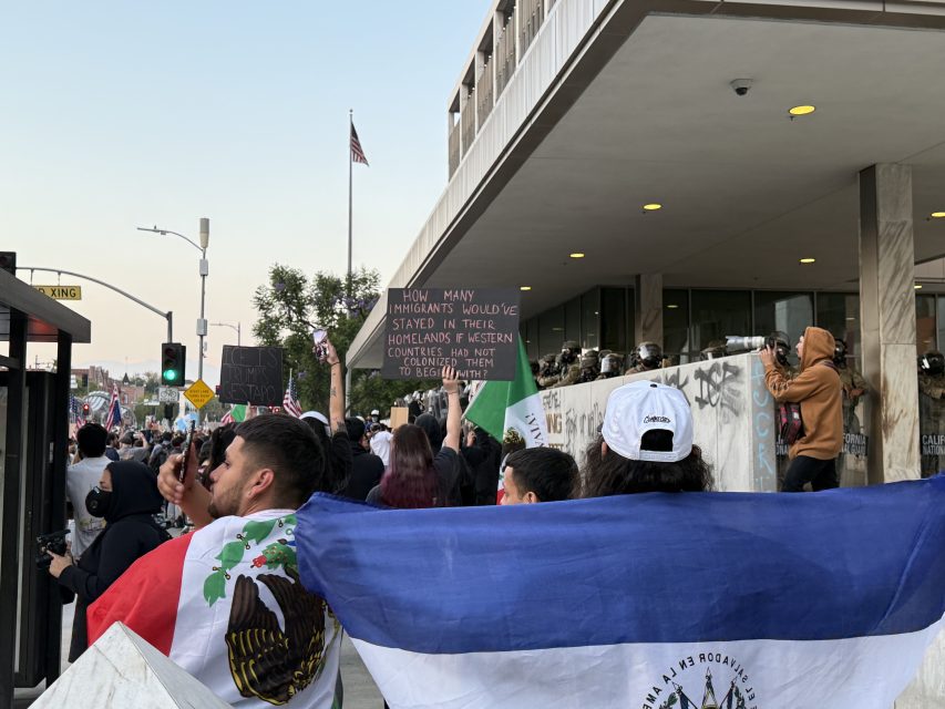A crowd of protesters holding flags and signs stands outside a building with graffiti, while some take photos; the American flag is visible in the background.