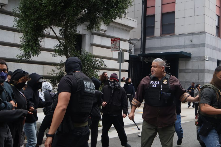 Police officers wearing "Federal Police" and "ICE" vests stand near a group of people, some in black clothing and masks, outside a city building.