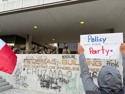 A protester holding a "Policy over Party" sign faces armed officers outside a graffiti-covered federal building in Los Angeles.