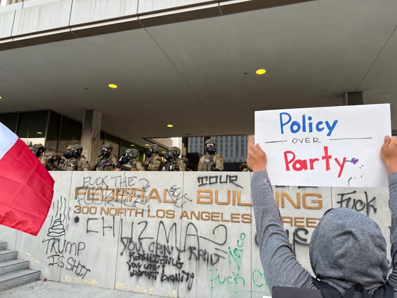 A protester holding a "Policy over Party" sign faces armed officers outside a graffiti-covered federal building in Los Angeles.