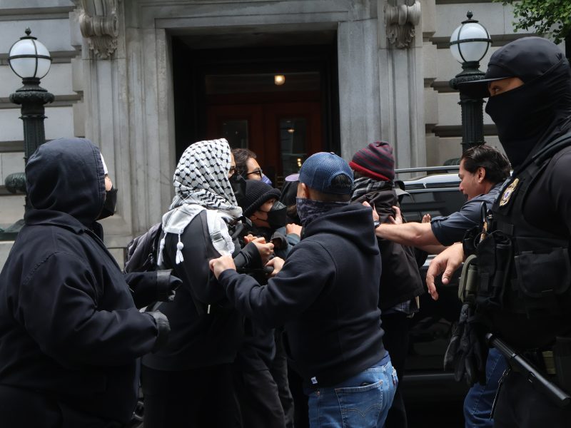 A group of people, some wearing masks and tactical gear, are involved in a physical altercation on a city sidewalk near a building entrance.