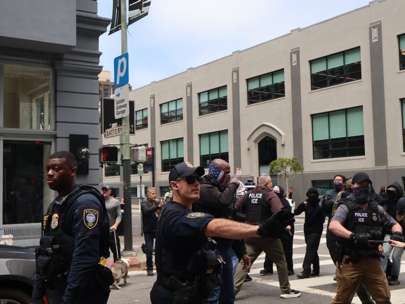 Police officers, some wearing "POLICE ICE" vests, stand and gesture on a city street near a group of people and a crosswalk, with buildings and parked cars in the background.