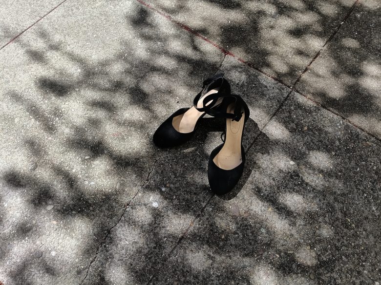 A pair of black closed-toe shoes with ankle straps sits on a sun-dappled concrete sidewalk.