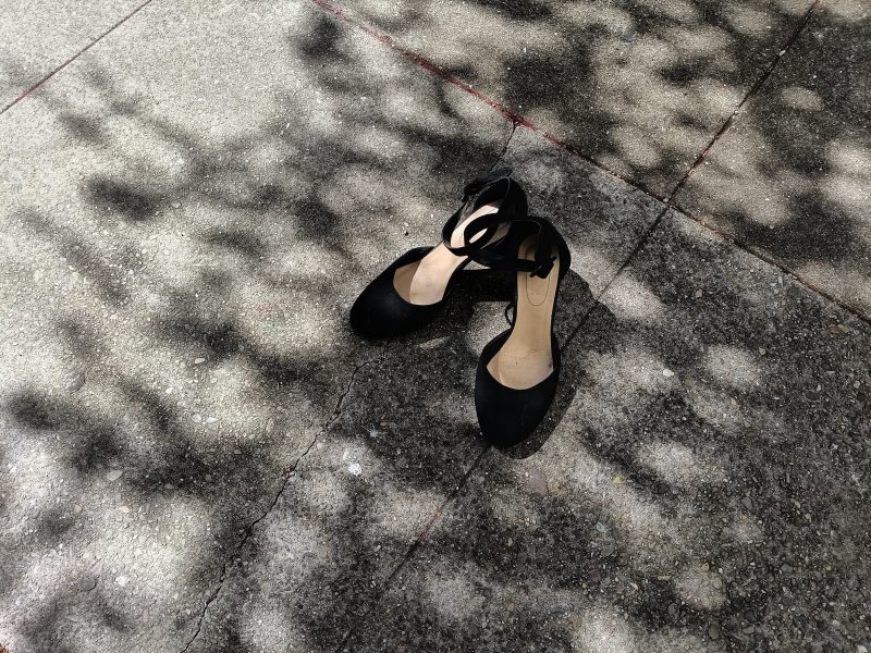 A pair of black closed-toe shoes with ankle straps sits on a sun-dappled concrete sidewalk.