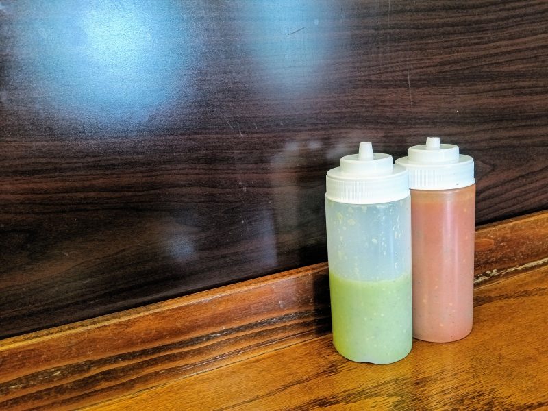 Two plastic squeeze bottles, one with green sauce and one with red sauce, are placed on a wooden surface against a dark wood-paneled background.