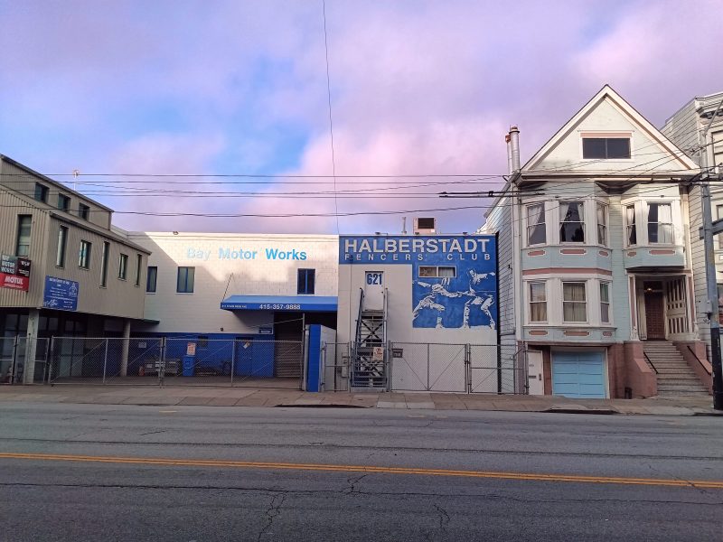 A street view of the Halberstadt Fencers' Club building with a mural, next to Bay Motor Works, under a cloudy sky.