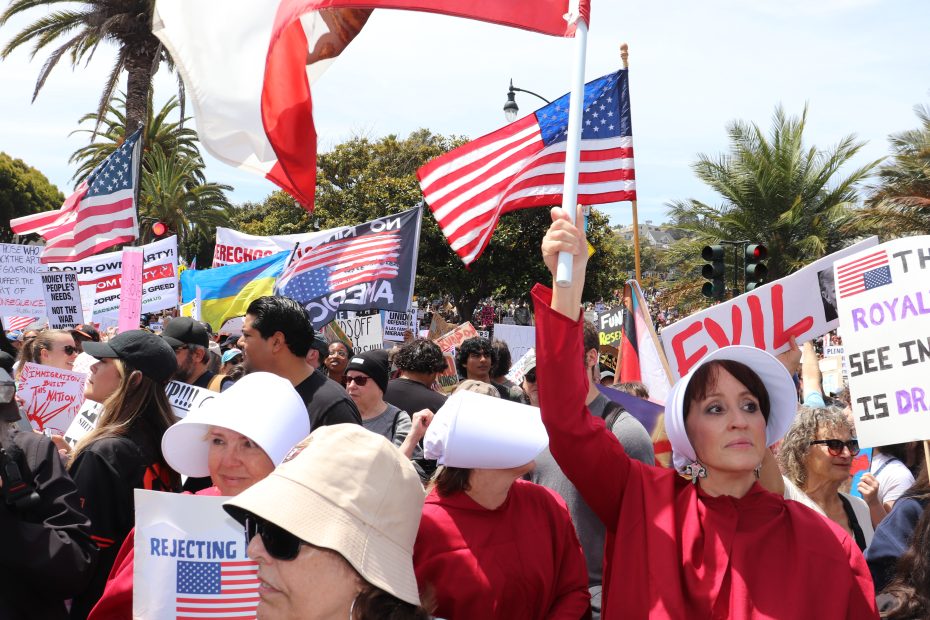 A group of protesters, some dressed in red Handmaid’s Tale costumes, hold protest signs and American flags at a demonstration outdoors.