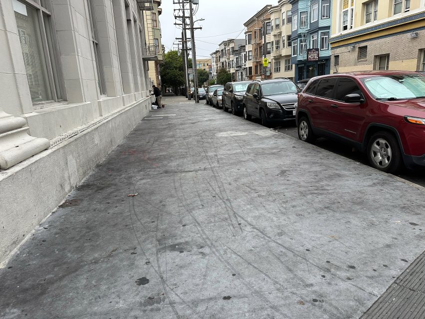 A city sidewalk with tire marks, parked cars along the street, and buildings lining both sides on an overcast day.