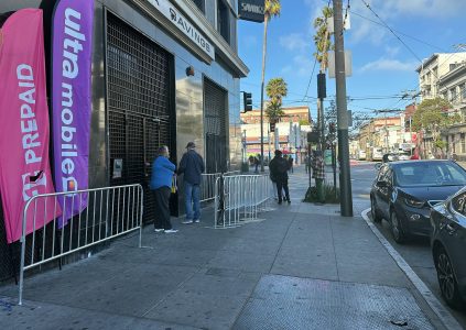Several people stand in line outside a store with "Ultra Mobile" and "Prepaid" banners on a city sidewalk, next to metal barricades and parked cars.