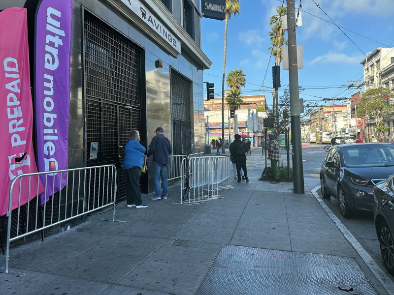 Several people stand in line outside a store with "Ultra Mobile" and "Prepaid" banners on a city sidewalk, next to metal barricades and parked cars.