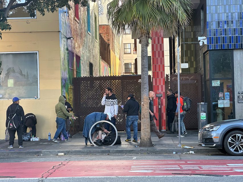 A group of people stand and sit on a city sidewalk near a building with colorful walls and a palm tree; some are gathered around a shopping cart.