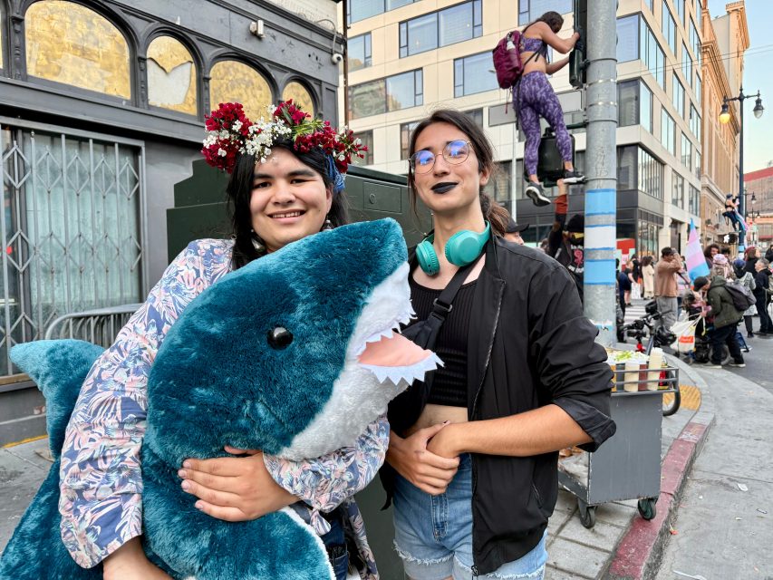 Two people pose on a city street during Pride; one holds a large plush shark and wears a flower crown, while the other sports headphones around their neck. People and buildings are visible in the background.