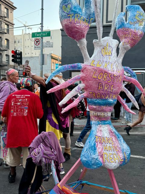 A large papier-mâché lobster sculpture with "Lobsters Are Trans" and "Trans Hate Is Real" painted on it stands proudly on a city street during a Pride event, surrounded by people showing their support.