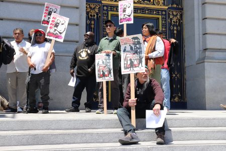 A group of people stand and sit on building steps holding protest signs with images and text; ornate doors are visible in the background.