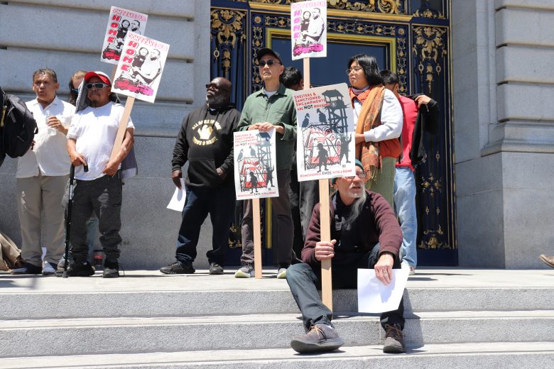 A group of people stand and sit on building steps holding protest signs with images and text; ornate doors are visible in the background.