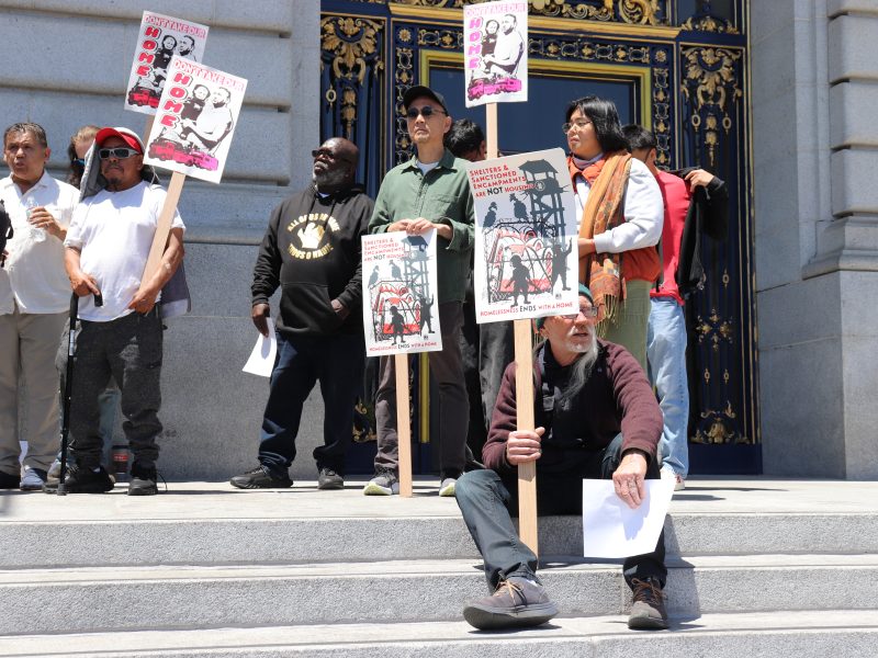 A group of people stand and sit on building steps holding protest signs with images and text; ornate doors are visible in the background.