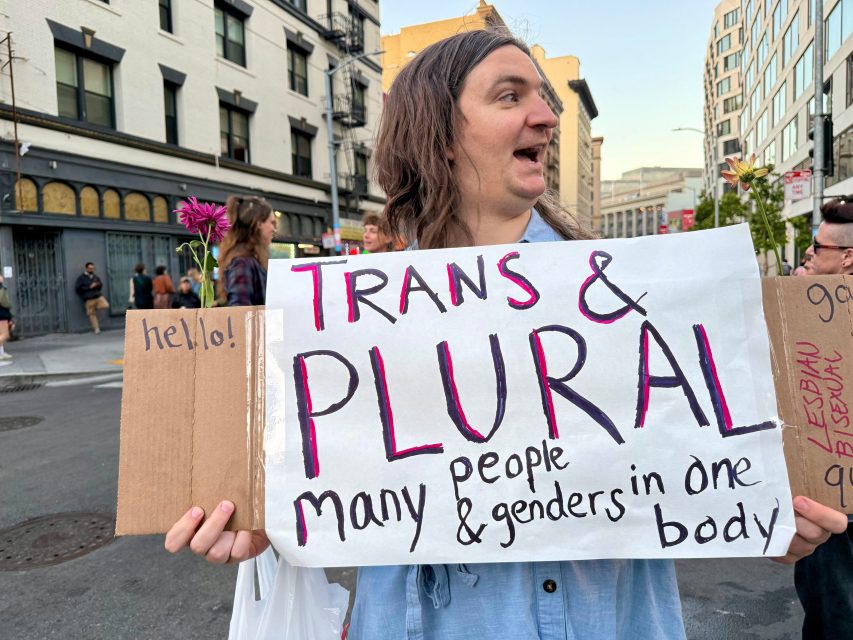 Person at a Pride street protest holding a sign reading "Trans & Plural—many people & genders in one body," with cardboard pieces saying "hello!" and "lesbian.
