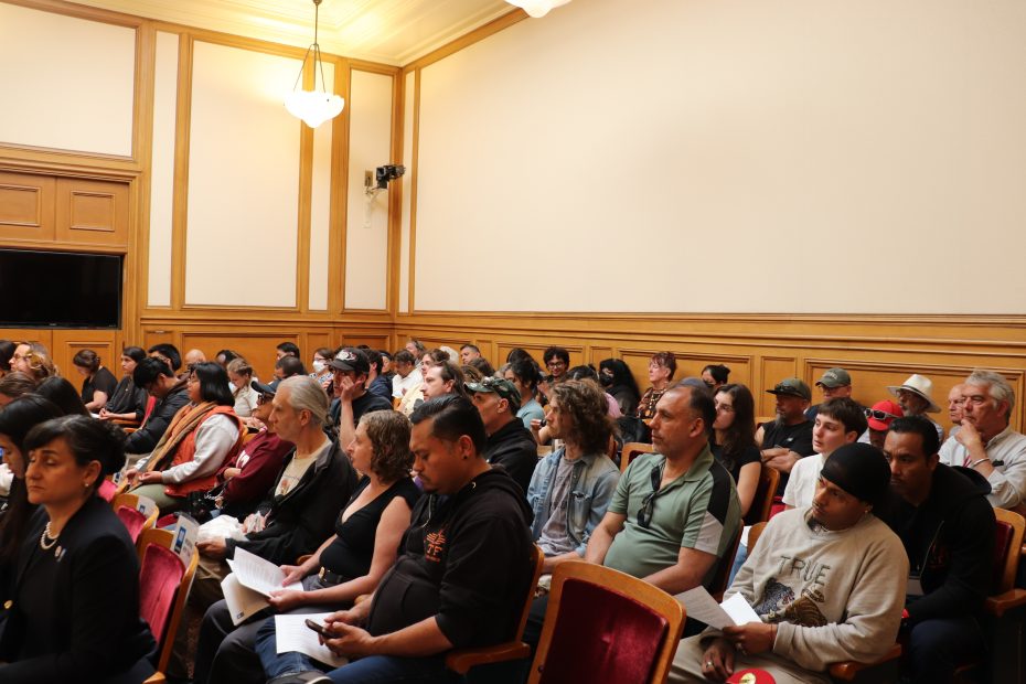 A diverse group of people sitting and listening attentively in a wood-paneled room, some holding papers, during a formal meeting or event.