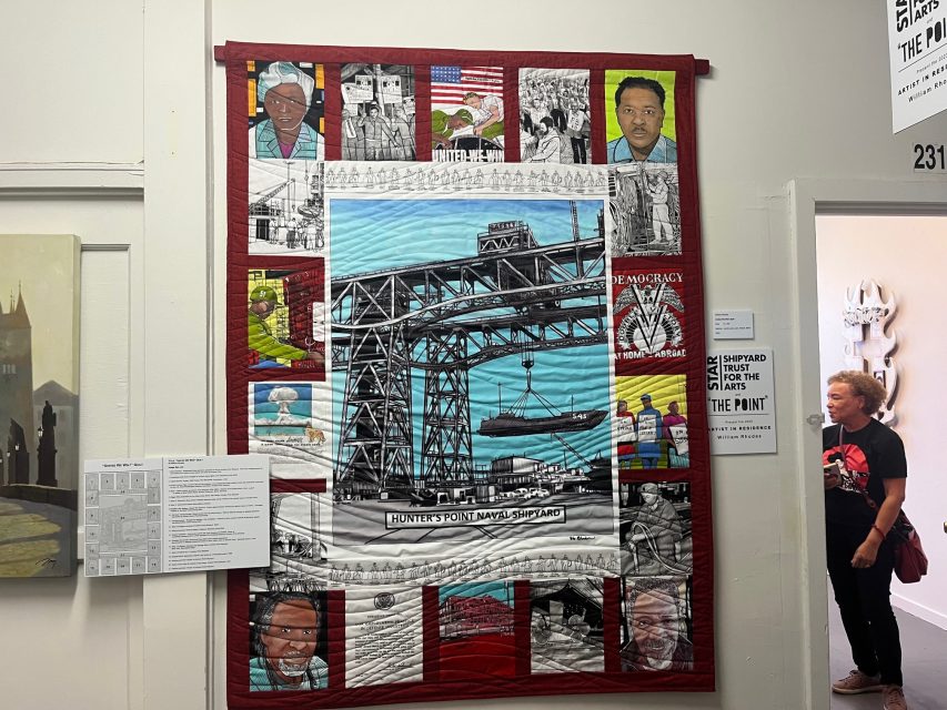 A colorful quilt hangs on a gallery wall, depicting the Hunters Point Naval Shipyard and portraits of people, with a visitor standing near the entryway.