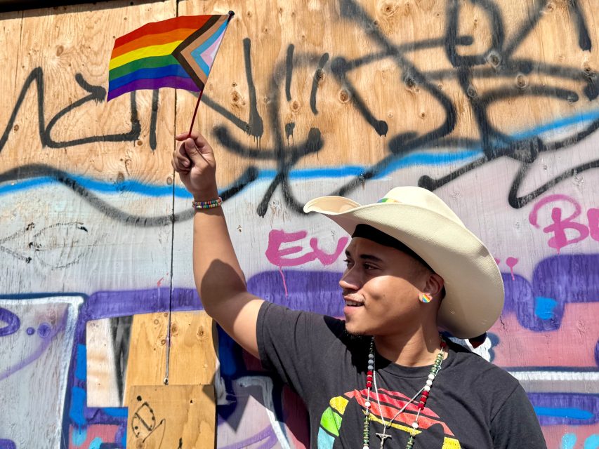 A person wearing a cowboy hat proudly holds up a Pride flag in front of a graffiti-covered wooden wall.