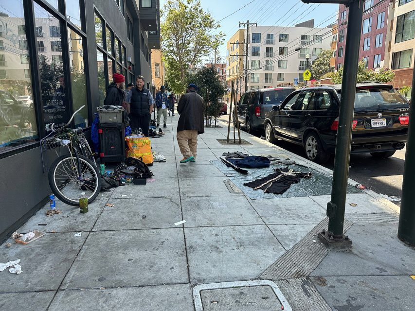 Several people stand and sit on a city sidewalk next to scattered belongings, a bicycle, and blankets, with parked cars and buildings in the background.