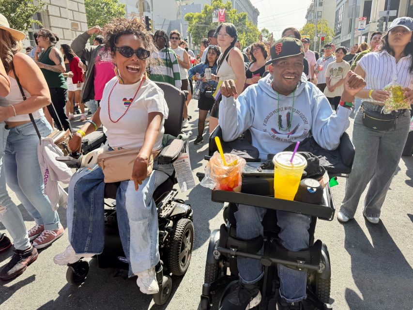 Two people in power wheelchairs smile and enjoy a lively outdoor Pride event, surrounded by a diverse crowd under sunny weather.