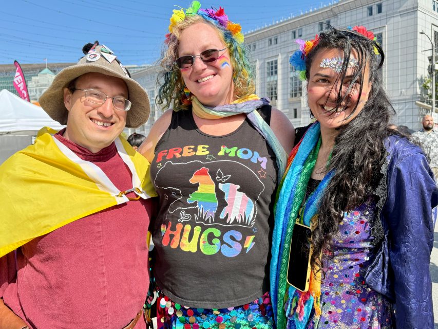 Three people smiling outdoors at a Pride event; one wears a "Free Mom Hugs" shirt, all sport colorful Pride clothing and accessories, with a building in the background.