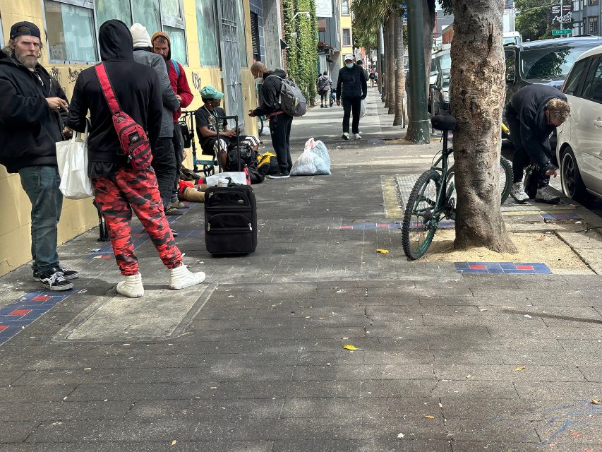 A group of people with bags and suitcases stand and sit along a city sidewalk, some near a tree and a bicycle, with buildings and parked cars in the background.