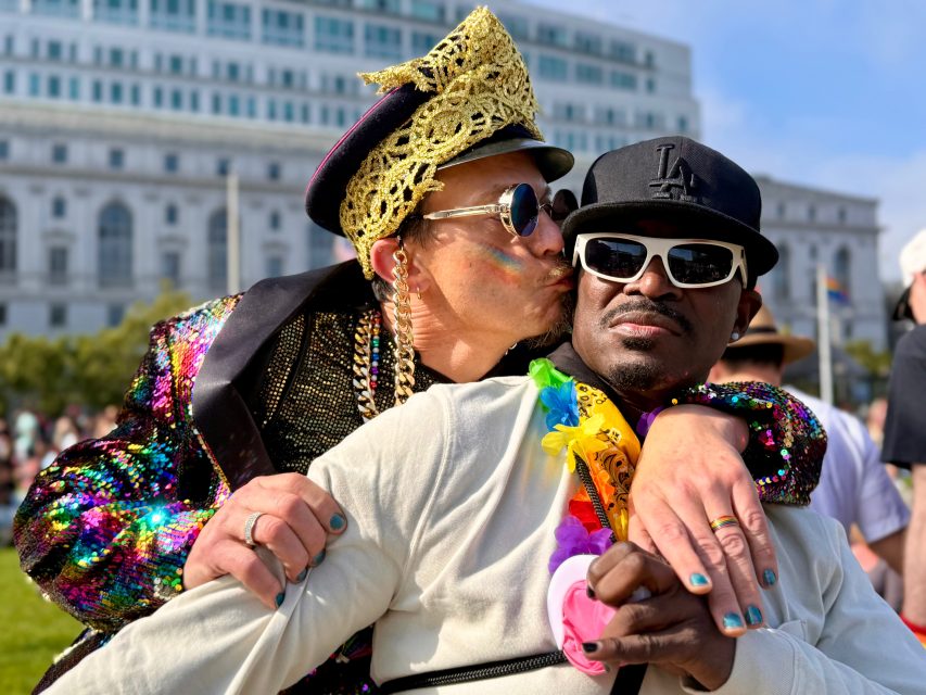Two men embrace at a Pride event; one in a colorful, sparkly outfit kisses the cheek of the other, who wears sunglasses, a black cap, and a white shirt with rainbow accessories.