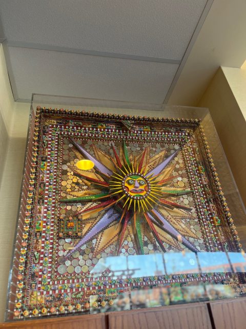 A square mosaic artwork featuring a colorful sun with a face at the center, surrounded by intricate patterns and small objects, displayed in a glass case.
