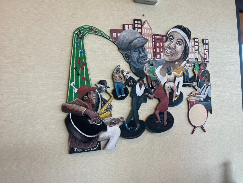 Wall art featuring musicians playing instruments, people dancing, and large faces in the background, set against a cityscape backdrop.