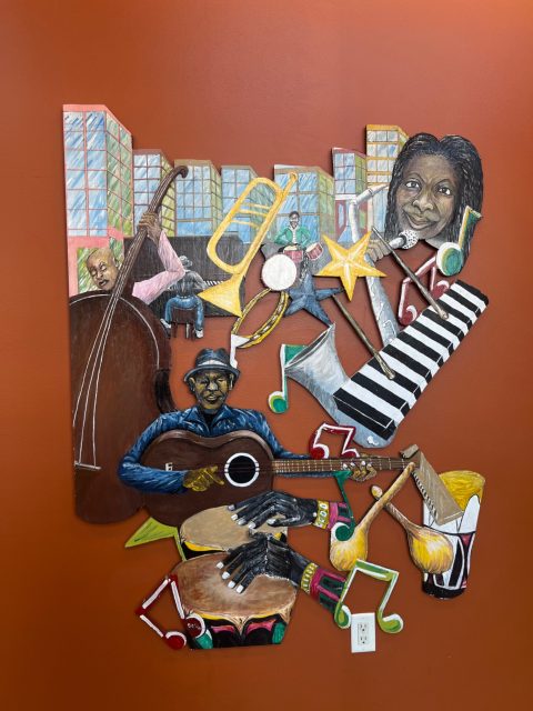 Colorful wall art depicting musicians playing instruments, musical notes, and city buildings on an orange background. A white electrical outlet is visible at the bottom center.