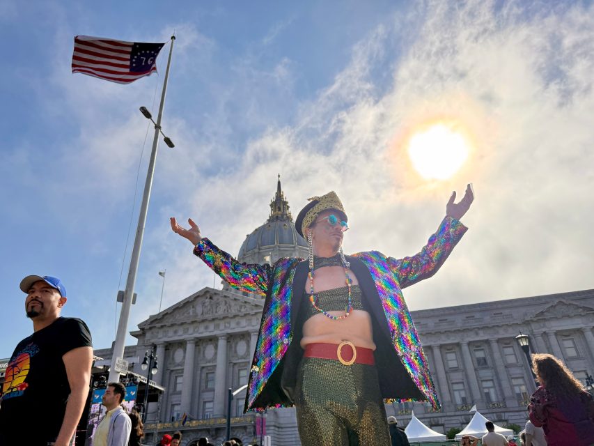 A person in a colorful sequin jacket and gold pants poses with arms outstretched, celebrating Pride in front of a domed building, with a U.S. flag and the sun shining in the background.