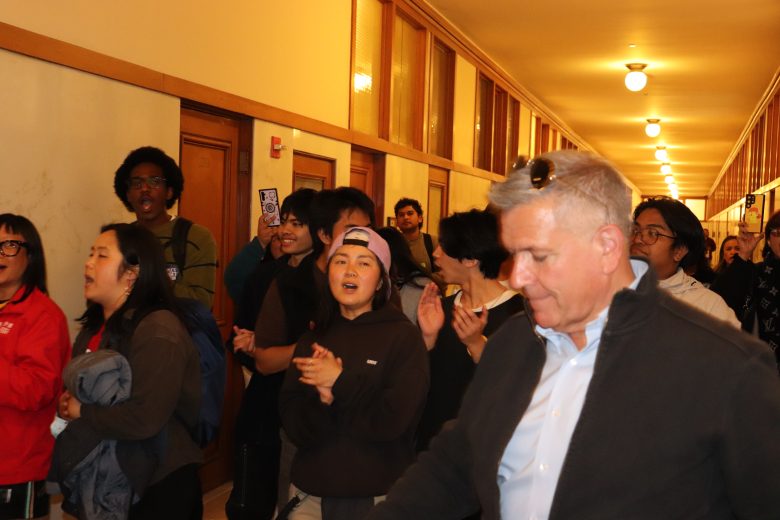 A group of people stand and clap in a hallway with wood-paneled walls and overhead lights. The focus is on a man in a jacket and sunglasses among others.