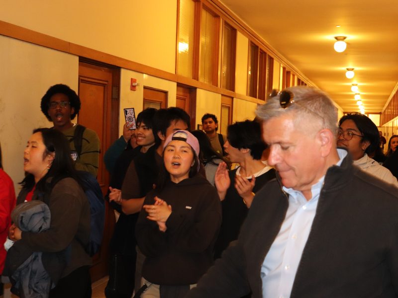 A group of people stand and clap in a hallway with wood-paneled walls and overhead lights. The focus is on a man in a jacket and sunglasses among others.