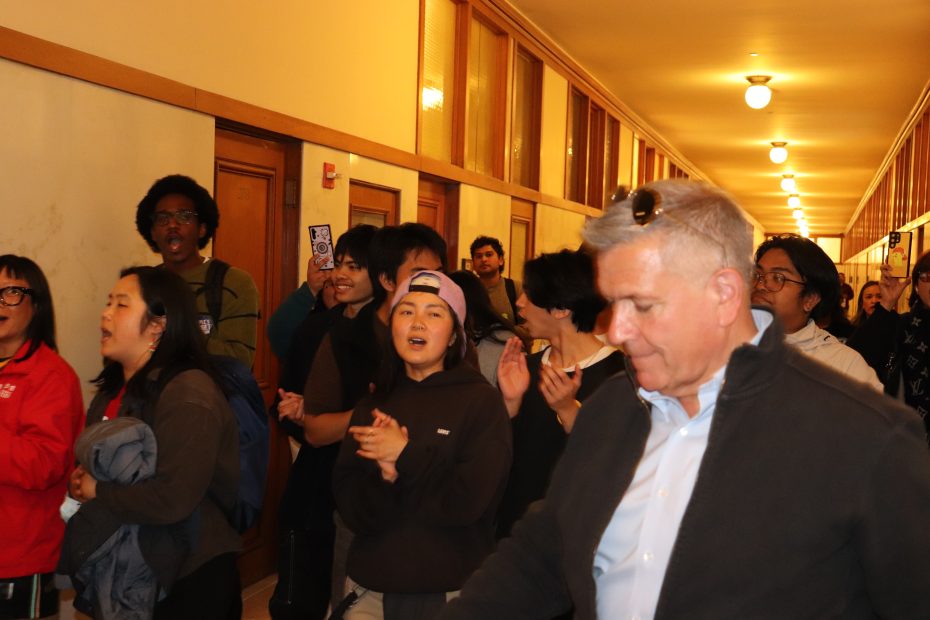 A group of people stand and clap in a hallway with wood-paneled walls and overhead lights. The focus is on a man in a jacket and sunglasses among others.