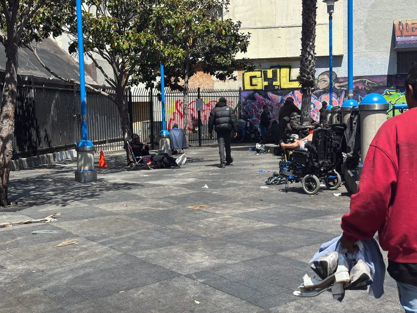 People are gathered in an urban outdoor area with some sitting, others standing, and graffiti on the wall in the background. A person in the foreground carries clothes.