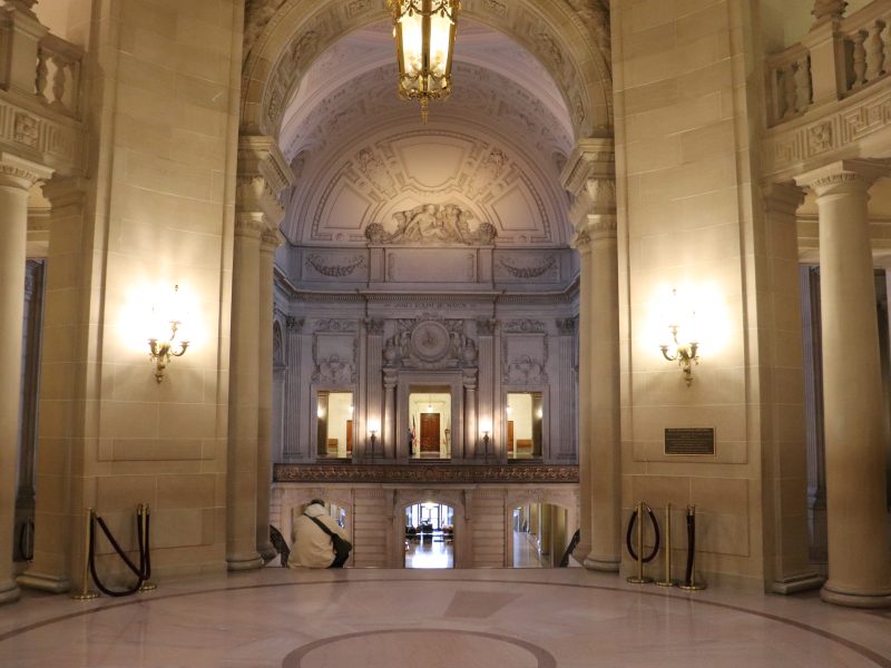 Grand interior hall with marble floors, ornate columns, arched ceiling, wall sconces, and a large chandelier in a historic building.