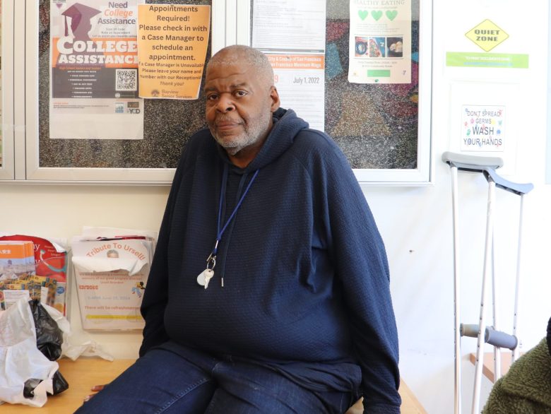 A man in a navy hoodie sits on a bench in front of a bulletin board filled with notices and balloons spelling "love" above him. A plant and crutches are nearby.